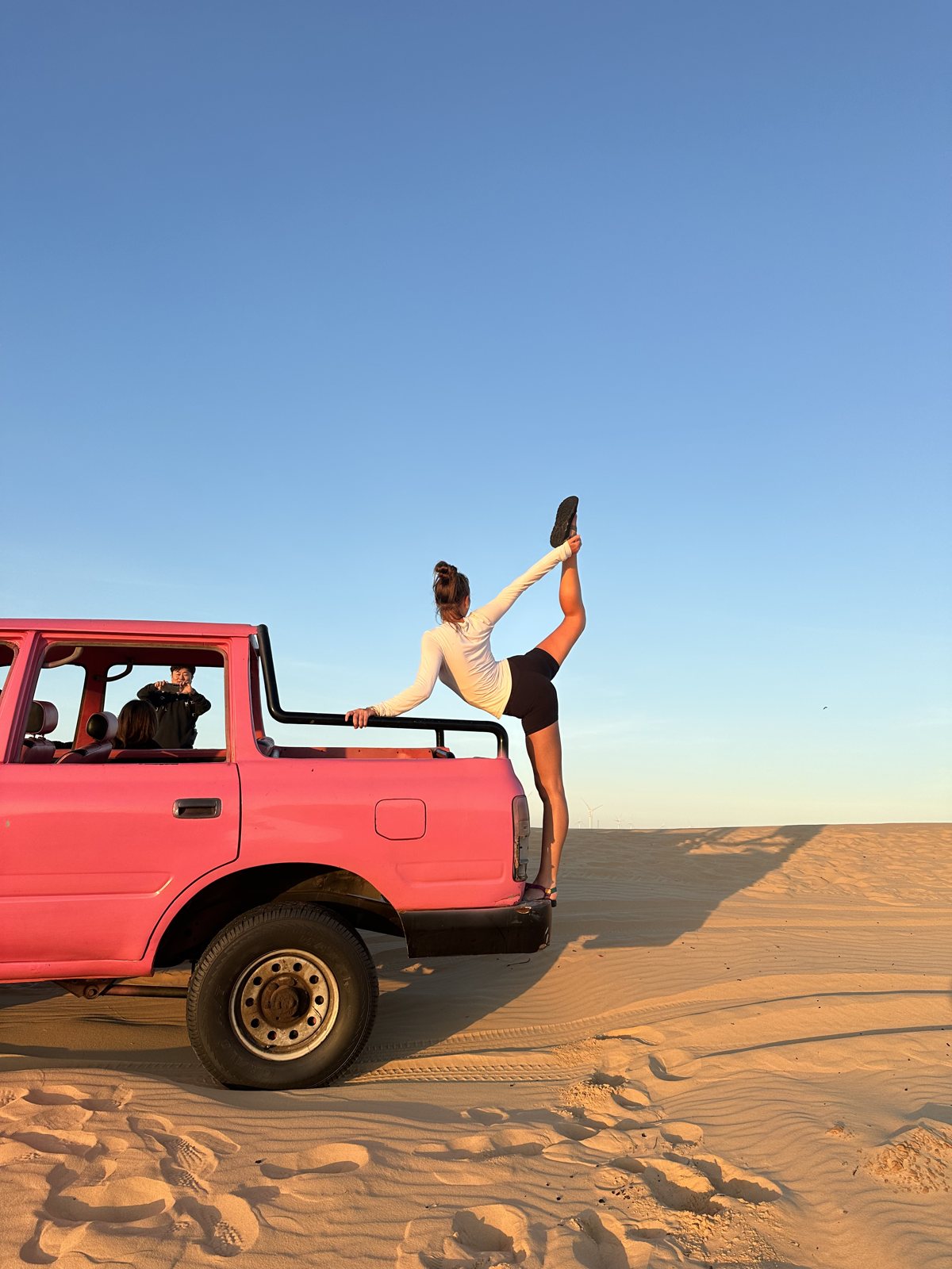 Hannah stretching against a pink truck on desert sand dunes